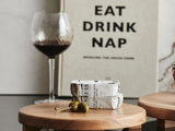 Et glas rødvin, stablede ostebåde og to oliven med tandstikkere på et træbræt. En bog med titlen EAT DRINK NAP ligger i baggrunden.