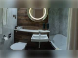 Moderne badeværelse med væghængt håndvask, toilet og badekar. Cirkulært spejl med belysning, træpaneler, marmorvægge og toiletartikler på disken.