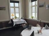 En hyggelig restaurant med hvide duge, sirligt foldede servietter, potteplanter, en lampe ved vinduet og et blødt naturligt lys, der kommer ind gennem de store vinduer på en tåget dag.