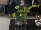 Tre glasvaser med grønne og hvide blomster på et sort sofabord foran en mørk sofa med guldben og en beige pude.