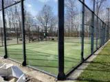 En padelbane med glasvægge og metalhegn, omgivet af nøgne træer og udendørs siddepladser. Himlen er klar, og jorden er dækket af grønt kunstgræs.
