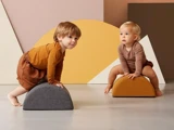 To småbørn i jordfarvet tøj leger med bløde, halvcirkelformede klodser i et moderne, minimalistisk rum med geometrisk vægkunst.