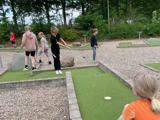 Børn spiller minigolf udendørs på en grøn bane omgivet af grus og træer. Et barn bøjer sig for at slå til en bold, mens andre ser på eller forbereder sig på at det bliver deres tur.