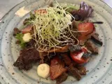 En farverig salat med spirer, cherrytomater, bladgrønt, mørke brødcroutoner og klatter af cremet dressing på en blå plettet tallerken.