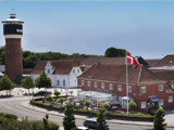 En rød murstensbygning med "Tønderhus" på forsiden, et vajende dansk flag og et højt muret vandtårn i baggrunden under en klar blå himmel.