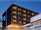 En moderne hotelbygning i skumringen, med varmt oplyste altaner og et lysende blåt HOTEL-skilt, der kan ses på den mørke facade.
