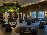 Elegant restaurantinteriør med runde borde, moderne stole, dæmpet belysning og et stort kunstigt træ i midten, som skaber en hyggelig og indbydende spisestemning.