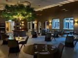 Moderne restaurantspisestue med runde borde, sorte stole, elegant borddækning og et stort kunstigt træ med lys i midten; varm belysning og store vinduer forbedrer stemningen.