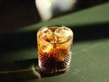 Et glas iskold mørk drik, muligvis whisky eller rom og cola, står på en grøn flade i sollys.