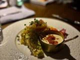 En anrettet gourmetret med en cremet dip i en lille messingskål, pyntet med mikrogrønt og sprøde toppings, serveret på en hvid tekstureret tallerken.