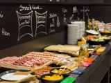 En morgenmadsbuffet med forskelligt pålæg, oste, brød i skiver, frugt, grøntsager og krydderier arrangeret på en disk foran en tavlemenu.