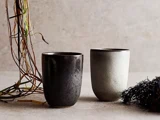 To keramiske kopper, en mørk og en lys, står på en struktureret overflade med tørrede kviste og blade i nærheden mod en neutral baggrund.