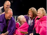 Fem mennesker, indhyllet i farverige tæpper, sidder sammen og smiler varmt, mens de rister pølser over et udendørs bål.