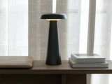 En moderne sort bordlampe står på en træplade ved siden af en lukket laptop og to stablede bøger med beige gardiner i baggrunden.