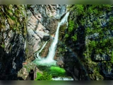 Et vandfald falder ned ad klipperne i en grøn pool med en svag regnbue synlig i tågen og den frodige grønne vegetation, der omgiver scenen.
