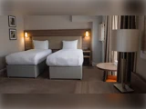 Et hotelværelse med to enkeltsenge, hvidt sengetøj, væghængte lamper, indrammet kunst, et rundt bord, en orange stol og et skrivebord med en lampe ved et vindue.