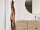 Et brunt håndklæde hænger på en krog ved siden af et badeværelsesbord i lyst træ med et rundt spejl over og små pyntegenstande på bordet.