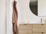 Et brunt håndklæde hænger på en knage ved siden af et rundt spejl over et badeværelsesbord i lyst træ med en vask og små pyntegenstande.