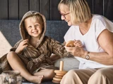 Et smilende barn i badekåbe sidder ved siden af en ældre kvinde med briller, og begge leger med trælegetøj, der kan stables, i en sofa.