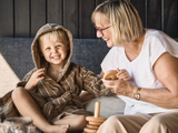 En smilende ældre kvinde med briller leger med en glad dreng i en brun badekåbe, begge sidder i en sofa med trælegetøj.