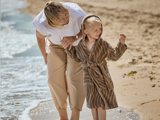 En voksen og et lille barn i morgenkåbe går langs stranden, holder hinanden i hånden og smiler, mens bølgerne rammer kysten.