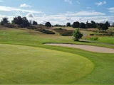 En velholdt golfbane med en sandbunker, grønt græs, spredte træer og en delvis overskyet blå himmel.