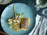 En anrettet gourmetret med stegt fisk med sprødt skind, grillede løg, en cremet sauce og garniture, serveret på en blå tallerken ved siden af et glas hvidvin og sølvtøj.