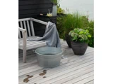 En metalspand står på en træterrasse ved siden af en bænk med et draperet håndklæde, en potteplante og spildt vand på dækket.