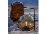 To moderne glaslamper på en quiltet overflade udendørs; den ene er en brun vaseformet lampe, den anden en rund ravfarvet lampe i en sort metalramme med en synlig pære indeni.