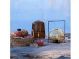 En picnic på et tæppe nær vand med brød, drikkevarer i glasflasker, en kurv, en lanterne og en brun glasvase.