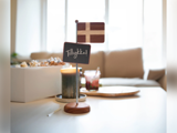 Et lille dansk flag og en kridttavle med Tillykke! på et stativ står på et bord i en lys, hyggelig stue.