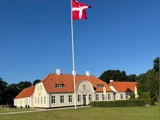 Et dansk flag vajer foran en stor cremefarvet bygning med rødt tegltag mod en klar blå himmel og omgivet af grønt græs og træer.