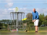 En mand i blå jakke kaster en gul disc mod en discgolfkurv markeret med tallet 3 på en græsklædt udendørs bane.