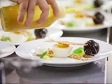 En hånd presser gul sauce ud over en anrettet gourmetdessert med en chokoladekugle, en cremet klat, en tynd, sprød skive og pynt på en hvid tallerken.