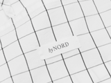 Nærbillede af et hvidt stof med et sort gittermønster og en påsyet etiket med byNORD i sort tekst.