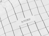 Nærbillede af et hvidt stof med et sort gittermønster og en påsyet etiket med byNORD i sort tekst.