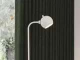 En moderne hvid gulvlampe med et rundt hoved står foran en mørk, lodret struktureret væg ved siden af et vindue med udsigt til træer udenfor.