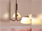Et nærbillede af en moderne lysekrone i messing med tre buede arme og kuppelskærme i matteret glas, der lyser blødt og varmt.