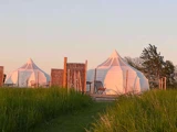 To hvide glampingtelte står midt i det høje græs ved solnedgang, med træhegn og udendørsstole i nærheden og træer i baggrunden.
