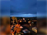 Folk spiser udendørs på en terrasse i skumringen, omgivet af stearinlys og dæmpet belysning, med en mørk, overskyet himmel og et åbent landskab i baggrunden.