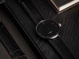 Et elegant sort armbåndsur med minimalistisk design og læderrem hviler på en mørk træoverflade ved siden af en bog.
