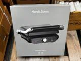 En kasse med en Nordic Sense Panini-grill på en træpalle, omgivet af andre kasser. Billedet af grillen og produktnavnet er trykt på forsiden af emballagen.