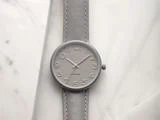Et minimalistisk gråt armbåndsur med store tal på et matchende gråt bånd, placeret på en lys marmoroverflade.