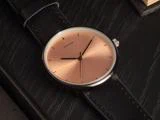 Et minimalistisk armbåndsur med en urskive i rosaguld, sorte timemarkeringer og en sort læderrem, der hviler på en mørk træoverflade.