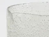 Nærbillede af en klar, struktureret glasbeholder med en bølget kant og hævede prikmønstre på en almindelig hvid baggrund.