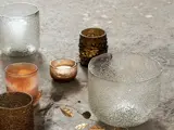 Flere teksturerede glaslysestager i forskellige størrelser og farver, bl.a. klar, guld og rav, er arrangeret på en stenoverflade med bladgulddekorationer.