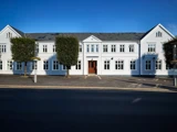 En stor hvid bygning med mange vinduer og en central trædør, flankeret af to grønne træer, under en klar blå himmel.
