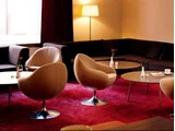 Moderne lounge med beige drejestole, lave borde med blomster, en mørk sektionssofa, rødt tæppe og en tændt gulvlampe i et svagt oplyst rum.