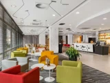 Moderne hotellobby med farverige lænestole, små runde borde, stribet gulvtæppe, et barområde i baggrunden og store vinduer med gennemsigtige gardiner i venstre side.