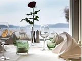 Elegant borddækning med foldede servietter, tomme vinglas, grønne glasskåle og en enkelt rød rose i en vase med en smuk udsigt over søen i baggrunden.